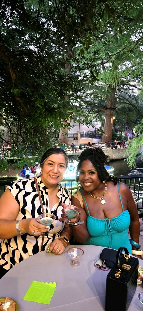 Top Shelf night: two guests with cocktails at a Riverwalk table under the trees